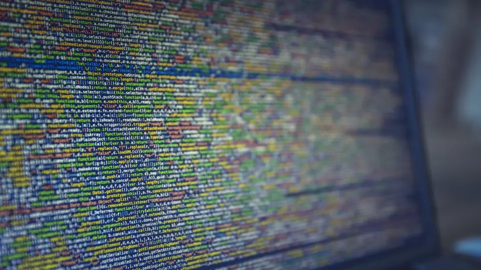 A laptop screen filled with dense minified code in blue, green and yellow, far too small to read line by line.