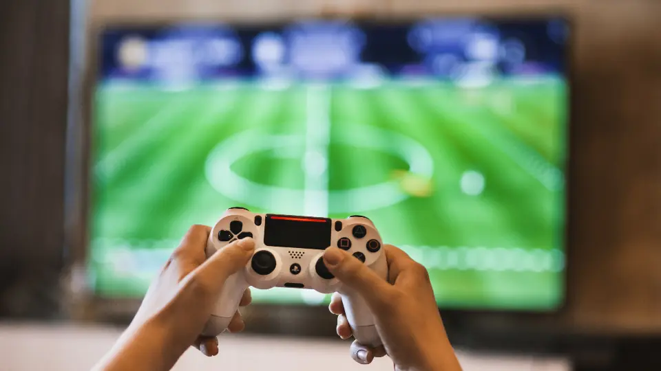 Two hands holding a white game controller in front of a television showing a football video game.