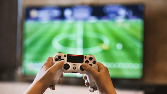 Two hands holding a white game controller in front of a television showing a football video game.