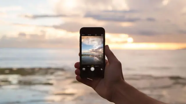 A hand holding a phone up against a sunset over the sea; the phone screen shows the same horizon it is framing.