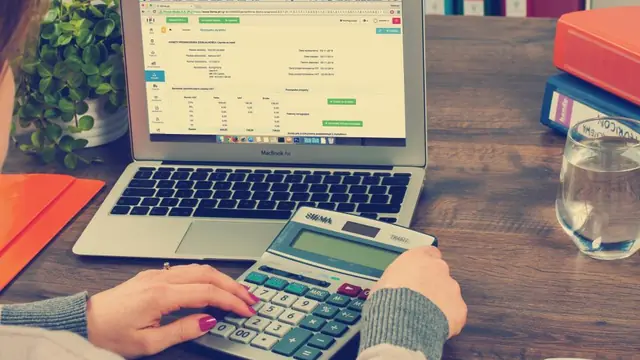 A person using a large desk calculator in front of a laptop that shows a filled-in accounting form.