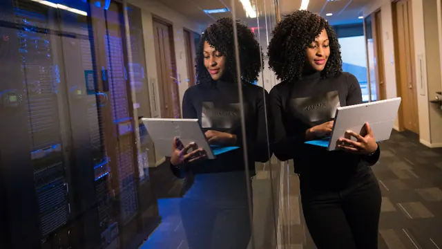 A woman holding an open laptop beside a glass wall of blue-lit server racks, her reflection in the glass.