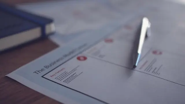 A printed worksheet with ruled boxes and small red headings lying on a wooden desk, a pen resting across it.