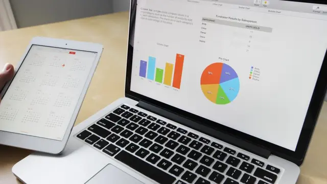 A laptop showing a spreadsheet with a small results table, a bar chart and a pie chart, beside a tablet held in one hand.