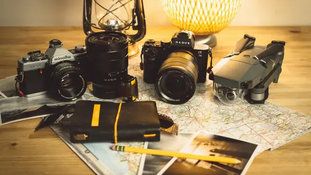 A film SLR, a mirrorless camera and a folding drone arranged on an open road map, with printed photos, a notebook and a lantern around them.