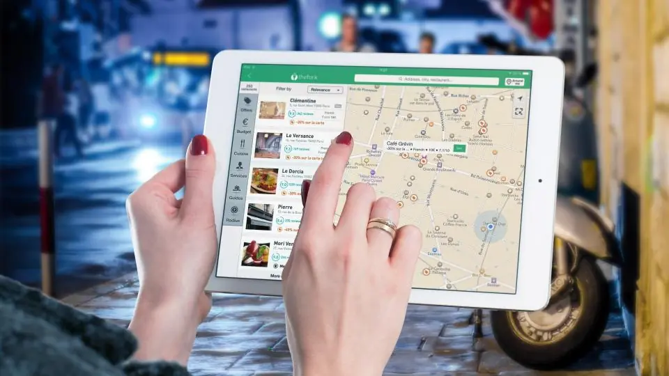Two hands holding a tablet that shows a city street map covered in location pins, on a wet street at night.