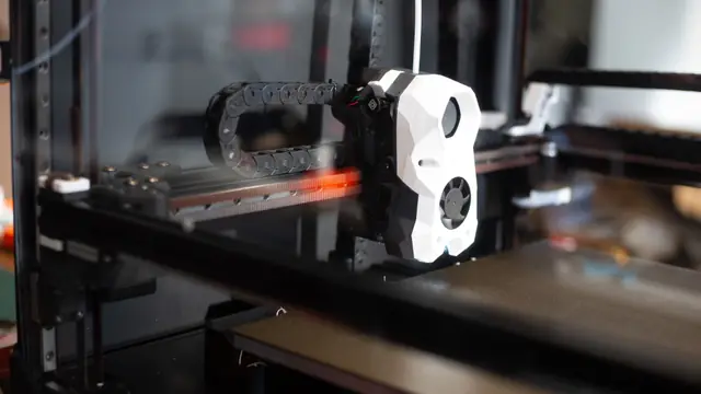The print head of a desktop 3D printer on its rail inside the frame, the drag chain behind it and an empty build plate below.