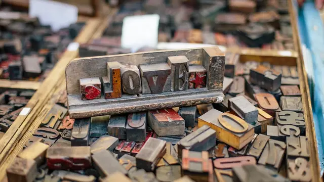 Wooden letterpress blocks spelling the word LOVE held in a metal frame, above a tray of mixed type.