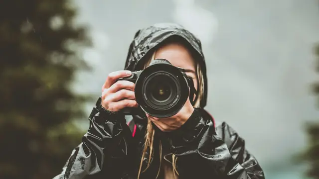 A photographer in a wet black rain jacket holding a DSLR up to her eye, blurred conifers behind her.