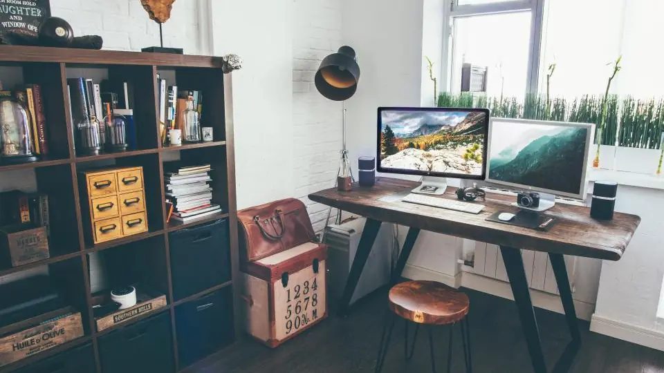A wooden desk by a window with two monitors side by side, each showing a mountain landscape photograph.