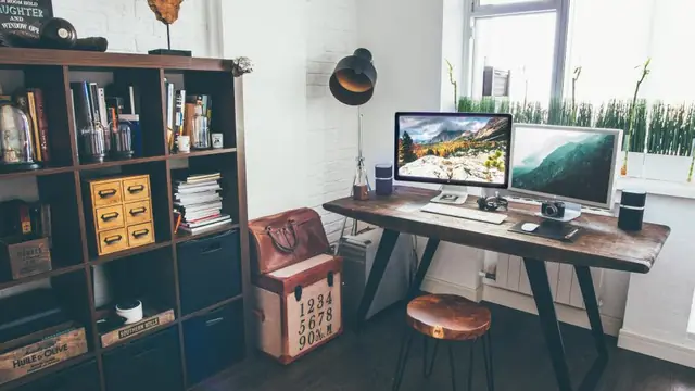A wooden desk under a window with two monitors side by side, each showing a landscape photograph.